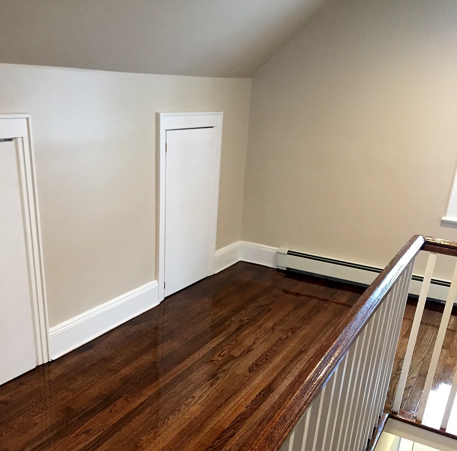 Upstairs hallway remodel in Baldwin, NY: a refinished brown wood floor along an upstairs hallway with a stained handrail and white spindles