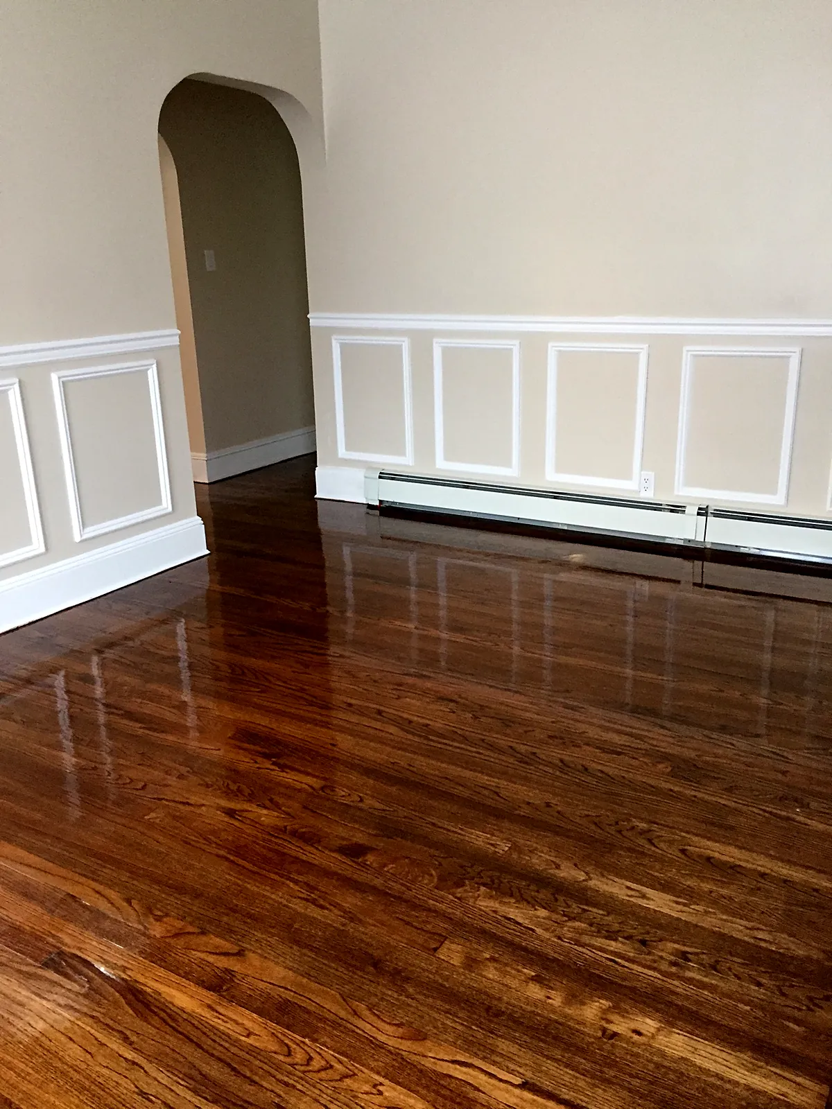 Living room remodel in Baldwin, NY: a refinished glossy brown wood floor in a room with white wainscot panels and an arched doorway