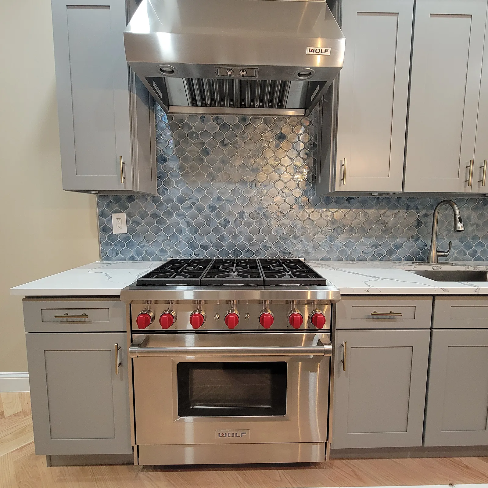 Kitchen remodel in the Bronx, NY: a stainless range and hood between gray shaker cabinets, with a blue mosaic backsplash and white counters