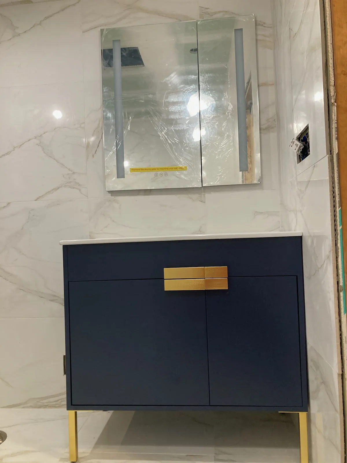 Bathroom remodel in Dix Hills, NY: navy vanity with gold legs, lit mirror cabinet and marble-look wall tile
