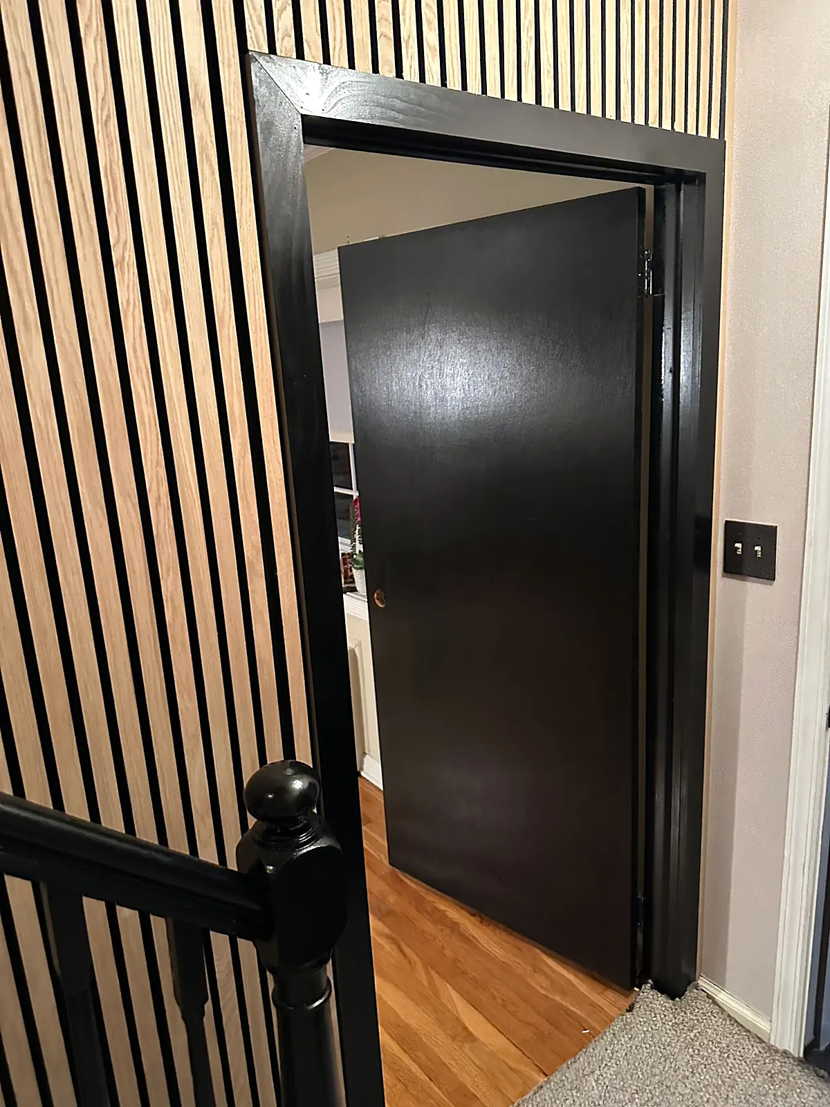 Stair landing remodel in Farmingdale, NY: a wood slat accent wall, a black-painted door and casing and a black newel post