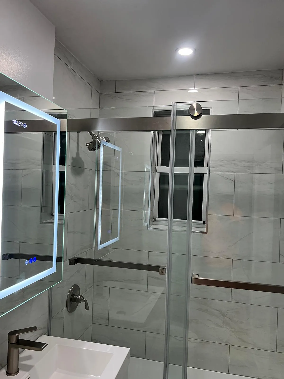 Bathroom remodel in Freeport, NY: a sliding glass tub door, large gray marble-look wall tile, a window and a lit mirror cabinet