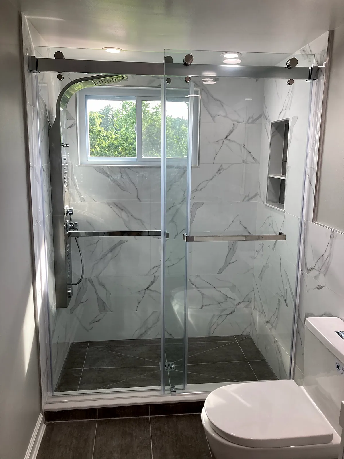 Bathroom remodel in Glen Head, NY: a glass sliding shower with marble-look walls, a tiled niche and a steel shower panel under a window