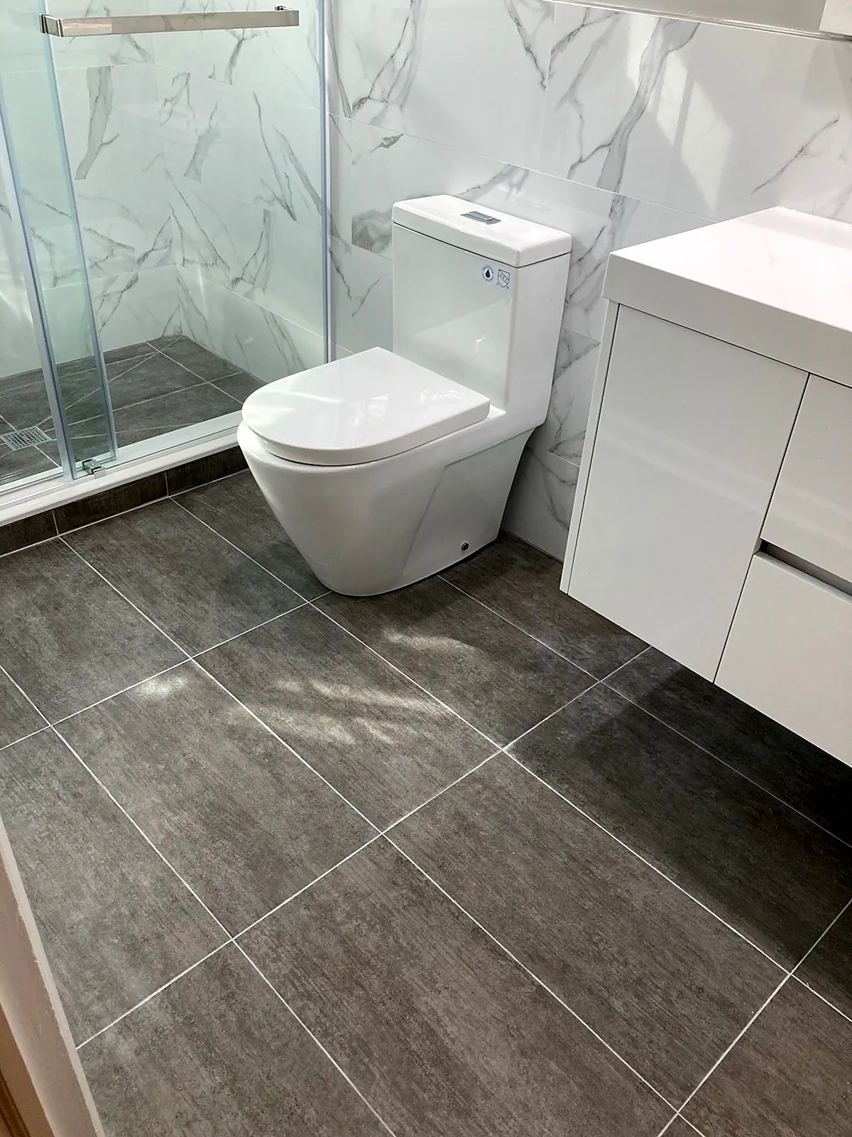 Bathroom remodel in Glen Head, NY: a one-piece toilet beside a white floating vanity on large gray floor tile, next to a glass shower