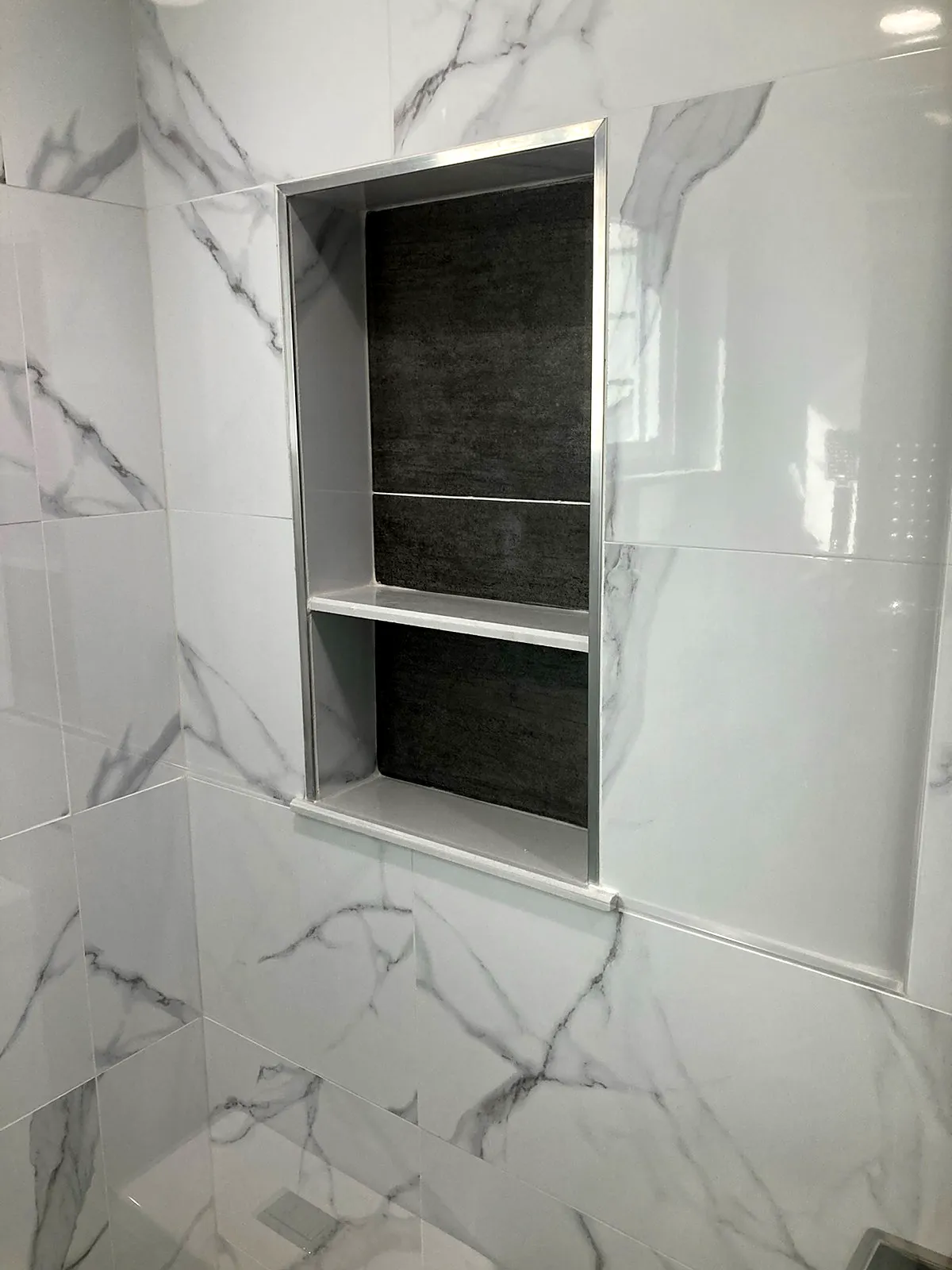 Shower remodel in Glen Head, NY: a recessed shower niche with a gray tile back and a shelf, framed by marble-look wall tile