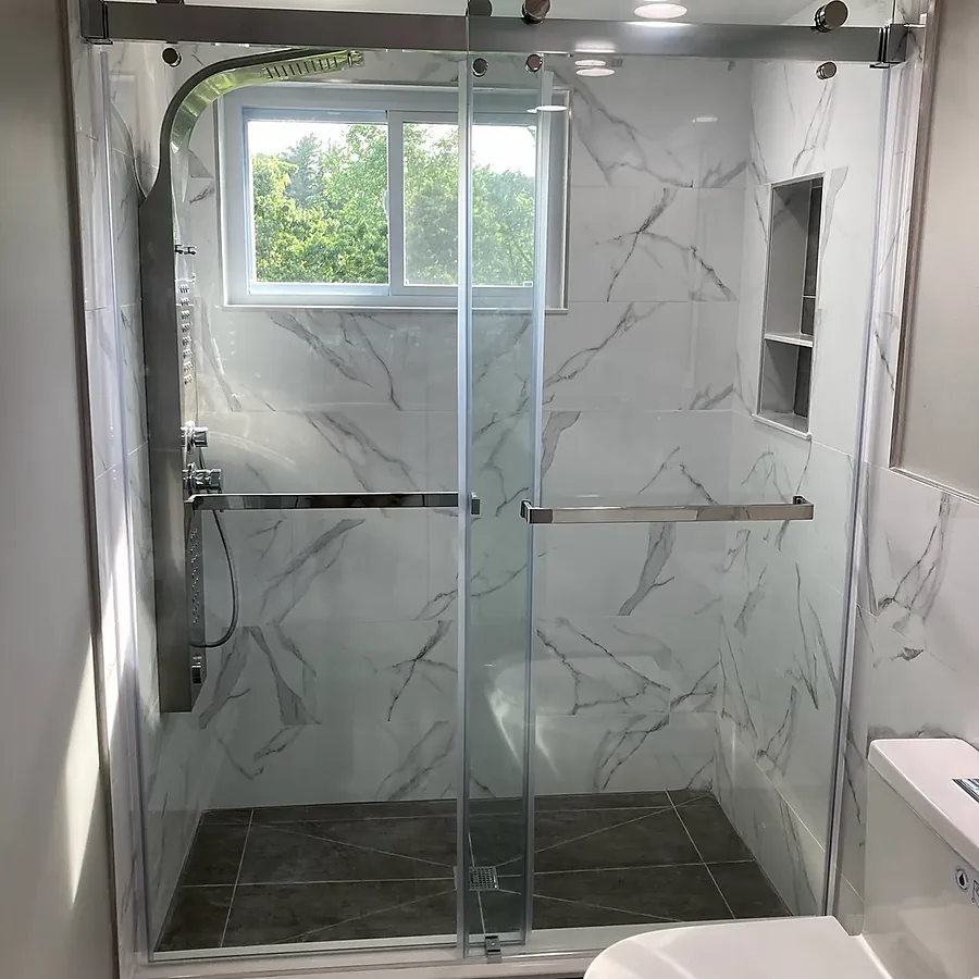 Bathroom remodel in Glen Head, NY: a glass sliding shower with marble-look walls, a tiled niche and a steel shower panel under a window