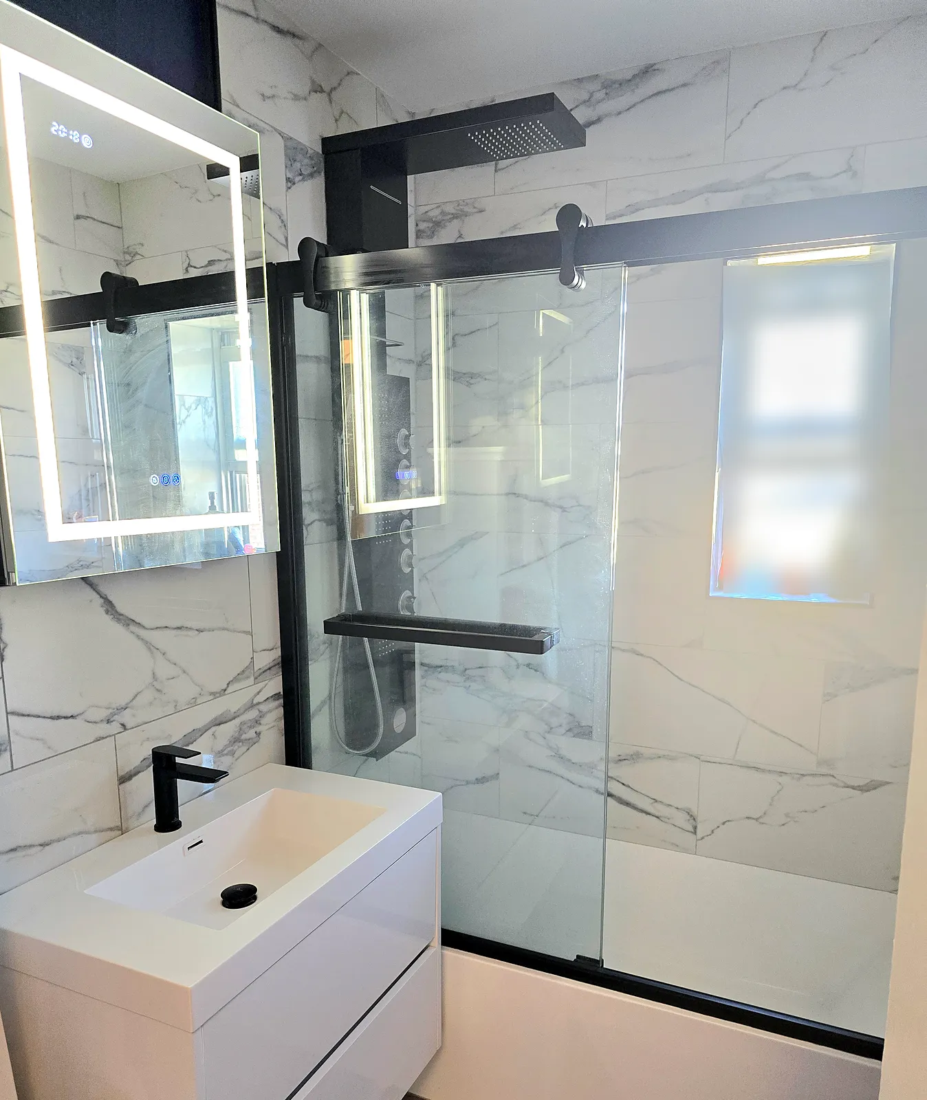 Bathroom remodel in Great Neck, NY: a white floating vanity, a lighted mirror and a black-framed glass tub door in marble-look tile
