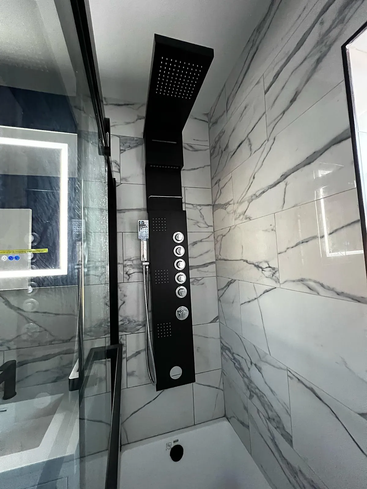 Bathroom remodel in Great Neck, NY: a black shower panel with body jets and a hand shower on marble-look tile over a white tub