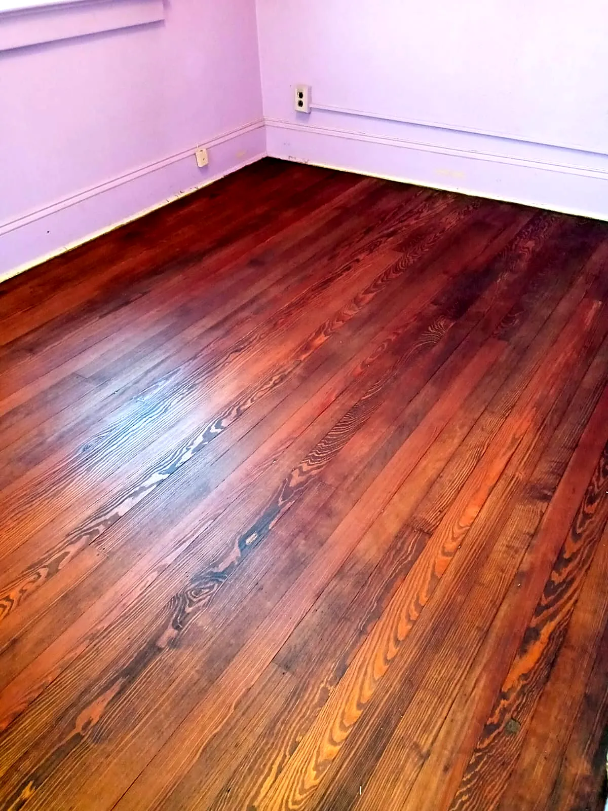 Bedroom remodel in Hempstead, NY: a refinished wide-grain wood floor with a warm reddish-brown stain in a lavender bedroom
