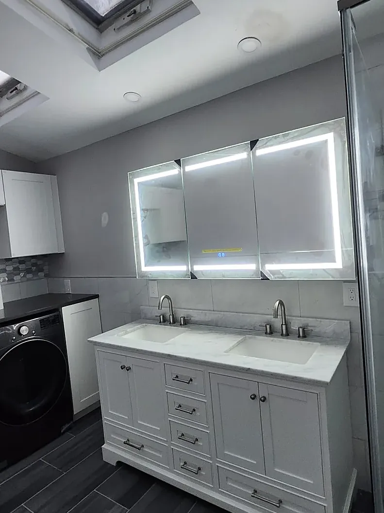 Bathroom remodel in Hicksville, NY: a white double vanity with a marble-look top and a wide lit mirror under two skylights, with dark plank-look tile