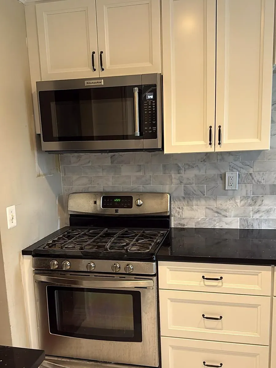 Kitchen remodel in Hicksville, NY: white raised-panel cabinets, a stainless range and microwave, a black counter and a marble-look tile backsplash