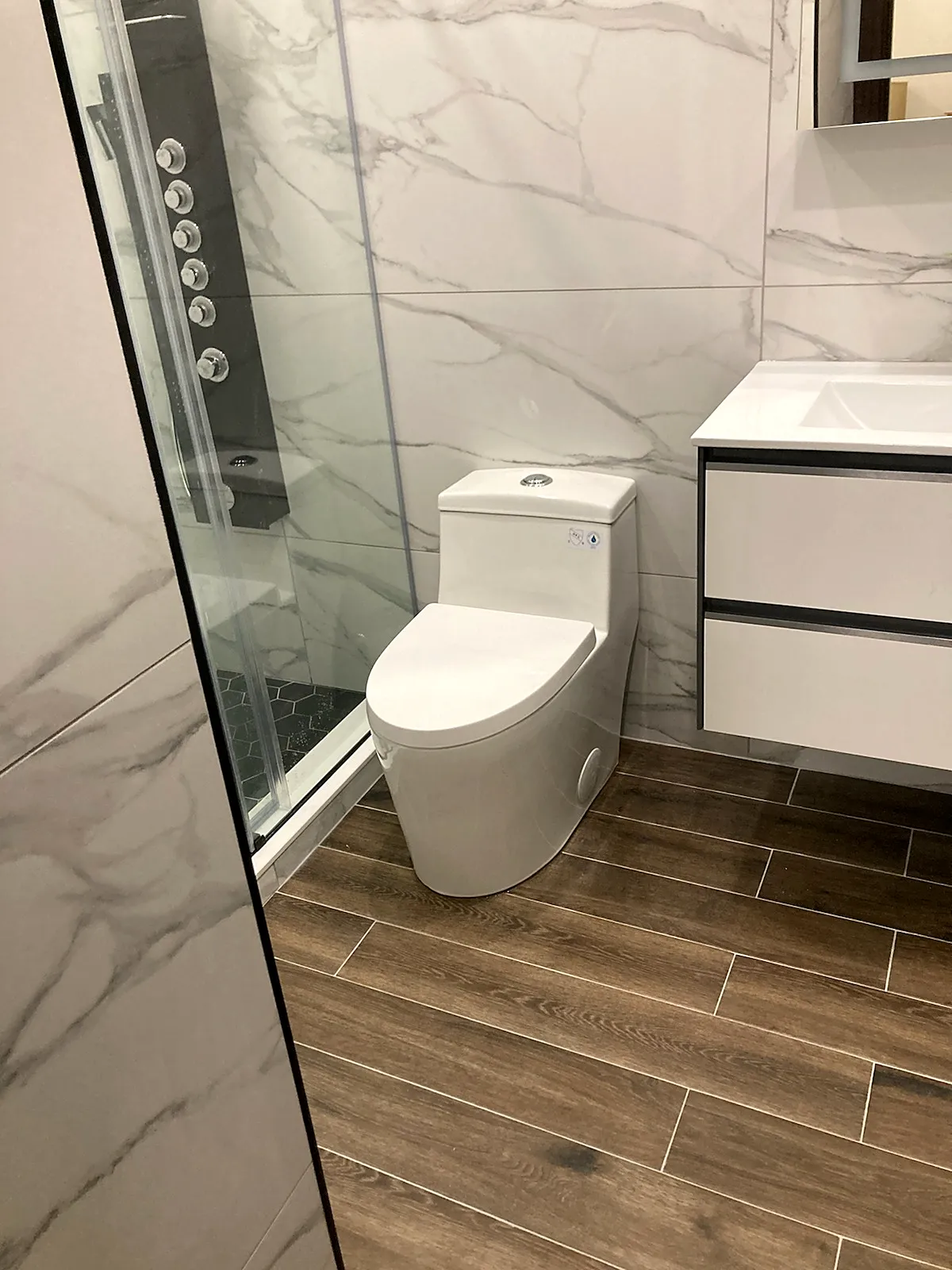 Bathroom remodel in Jericho, NY: a white floating vanity, a one-piece toilet and a glass shower with marble-look walls and a wood-look floor