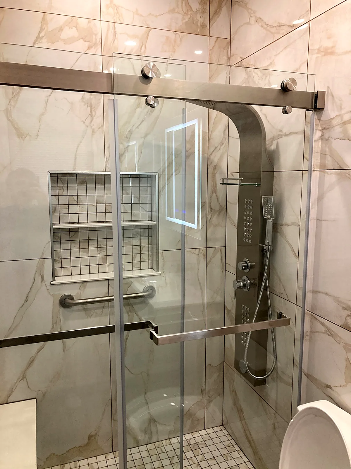 Bathroom remodel in Jericho, NY: a glass sliding shower with a steel shower panel, grab bars and a tiled niche in marble-look tile