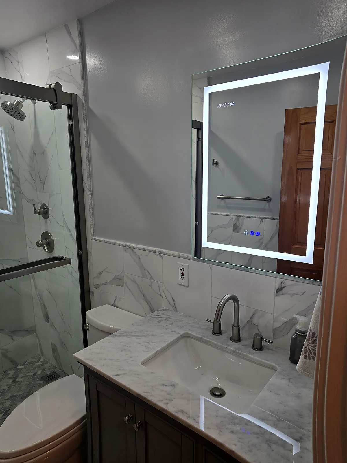 Bathroom remodel in Levittown, NY: a dark vanity with a white marble-look top, a lit mirror and marble-look wall tile beside a glass shower