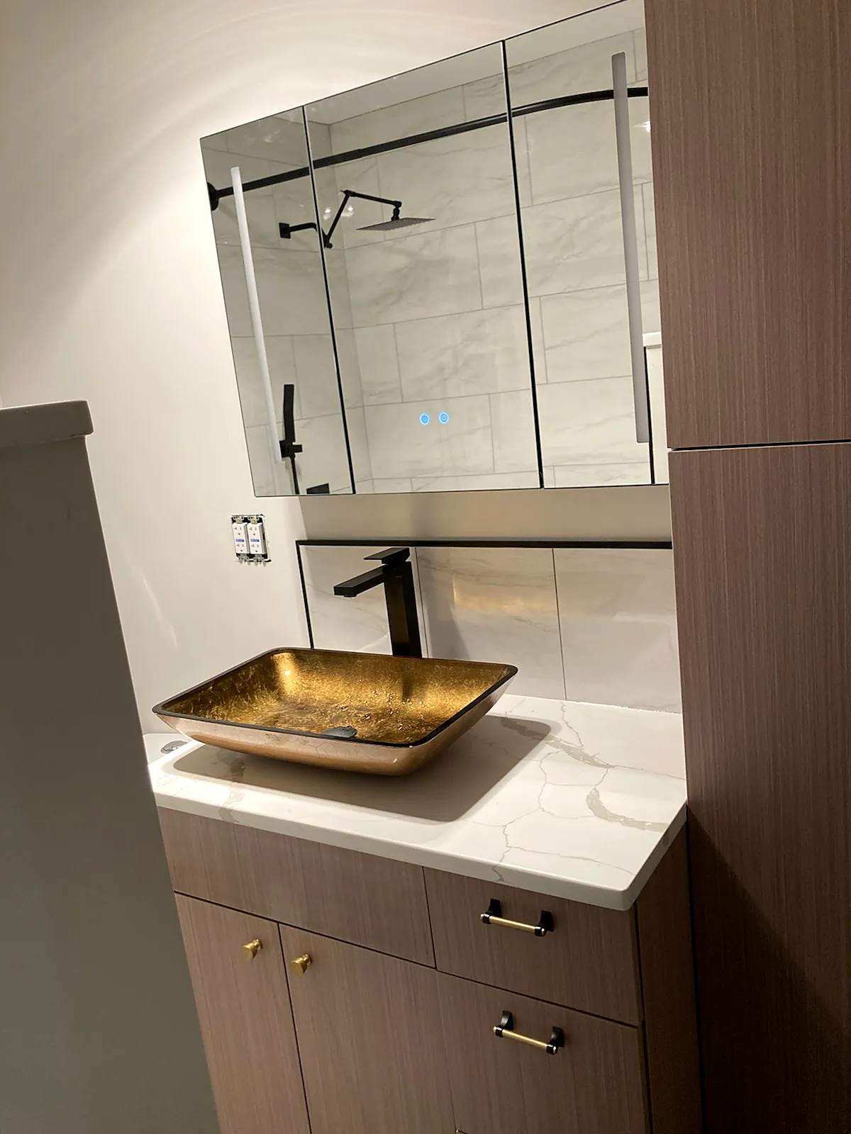 Bathroom remodel in Lindenhurst, NY: a wood-look vanity with a gold glass vessel sink, a lighted mirror cabinet and a tall linen cabinet