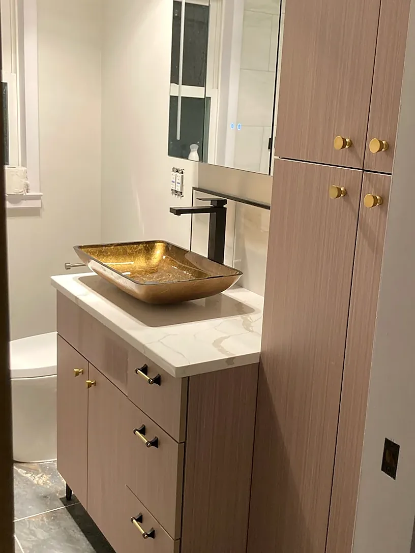 Bathroom remodel in Lindenhurst, NY: a wood-look vanity and linen tower with brass knobs, a gold vessel sink and dark stone-look floor tile