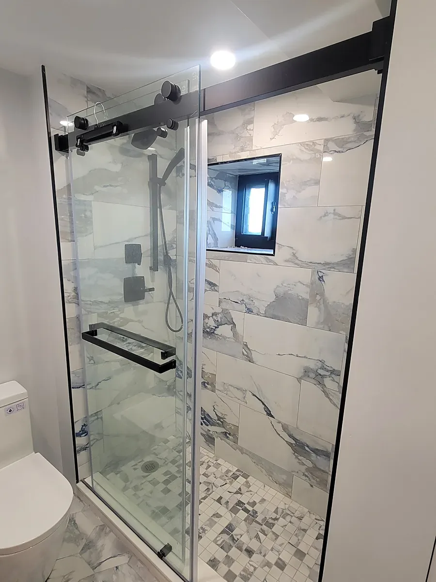 Bathroom remodel in Long Beach, NY: a sliding glass shower with black hardware, marble-look wall tile, a mosaic shower floor and a window