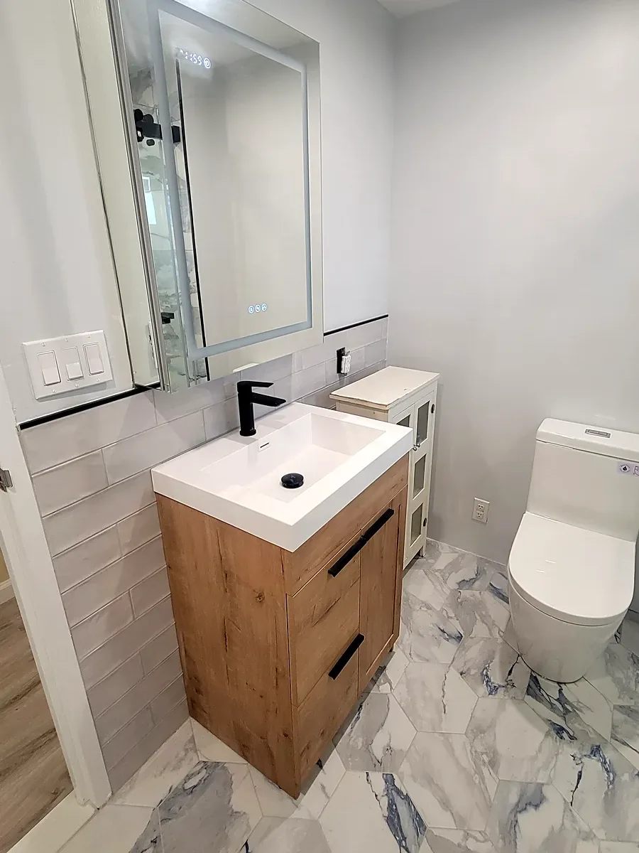 Bathroom remodel in Long Beach, NY: a wood vanity with a white sink, a lit mirror cabinet and blue-veined marble-look hexagon floor tile