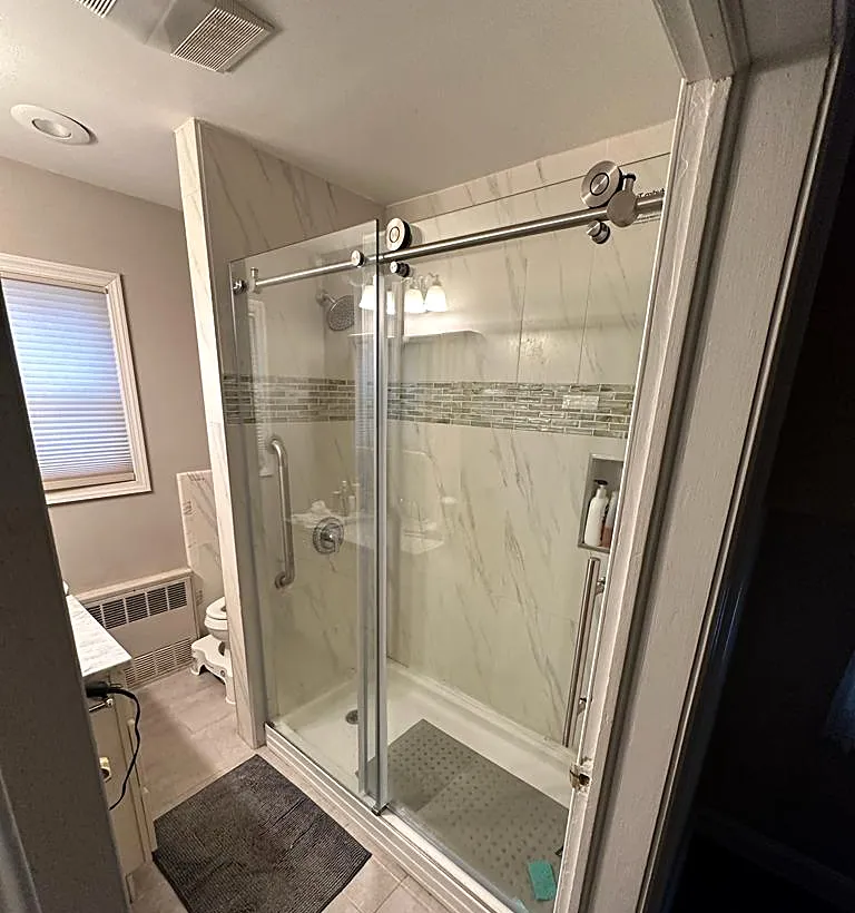 Bathroom remodel in Lynbrook, NY: a walk-in shower with a sliding glass door, marble-look walls and a glass mosaic band
