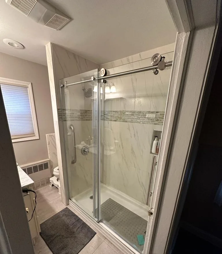 Bathroom remodel in Lynbrook, NY: a glass slider on a steel top bar in front of a walk-in shower, with gray floor tile and a window beyond