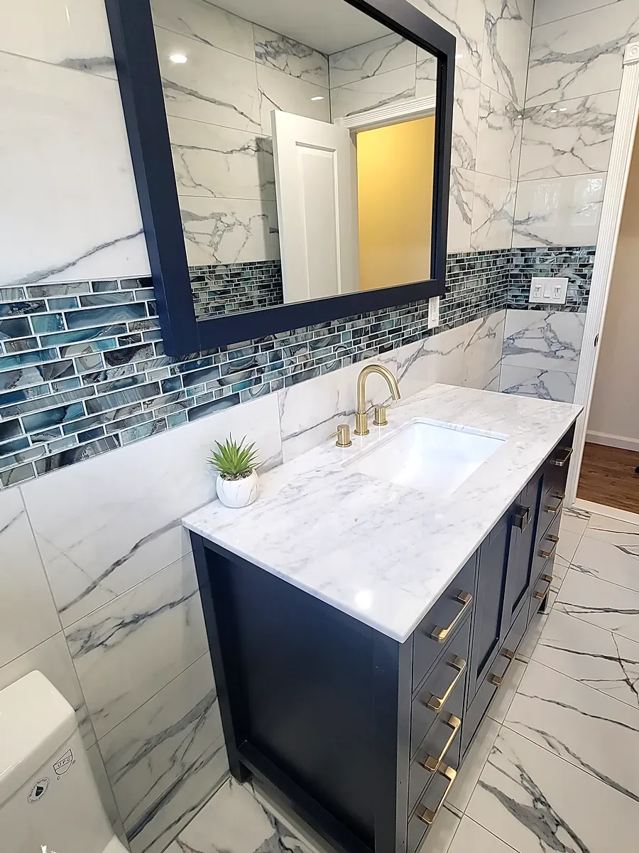Bathroom remodel in Middle Island, NY: a navy vanity with a marble-look top, gold faucet, navy-framed mirror and a blue glass tile band