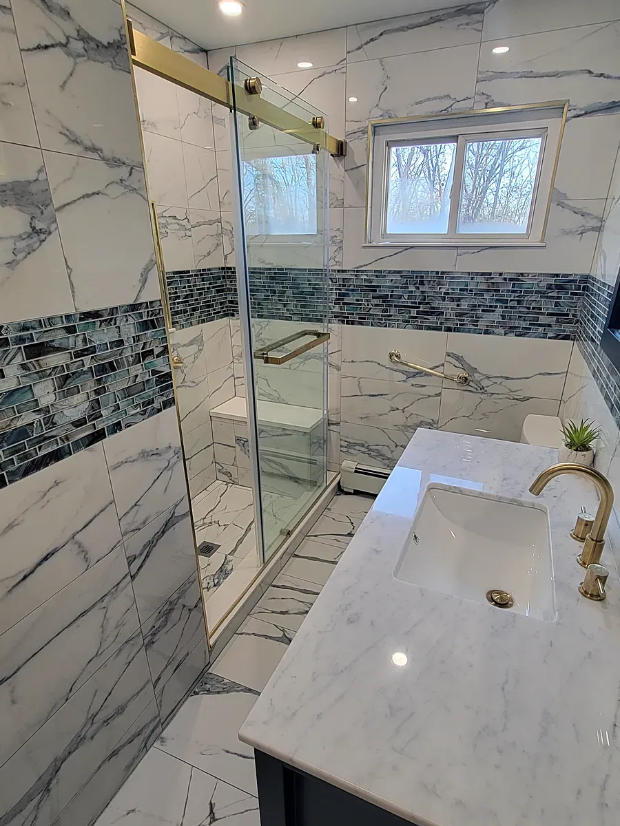 Bathroom remodel in Middle Island, NY: a navy vanity beside a gold-framed glass shower, with marble-look tile and a blue glass tile band