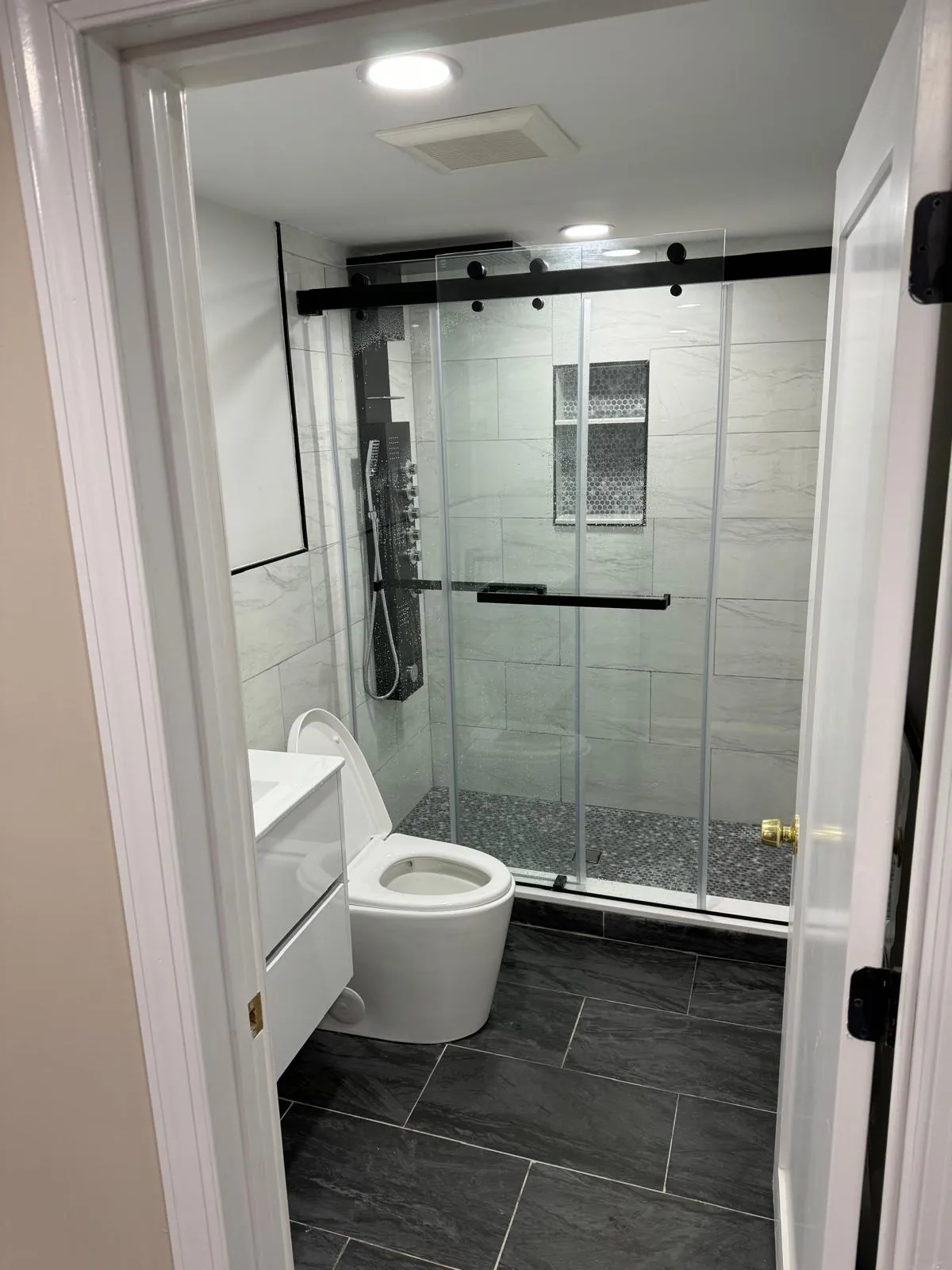 Bathroom remodel in New Hyde Park, NY: sliding glass shower with black hardware, gray tile, tiled niche and dark floor tile
