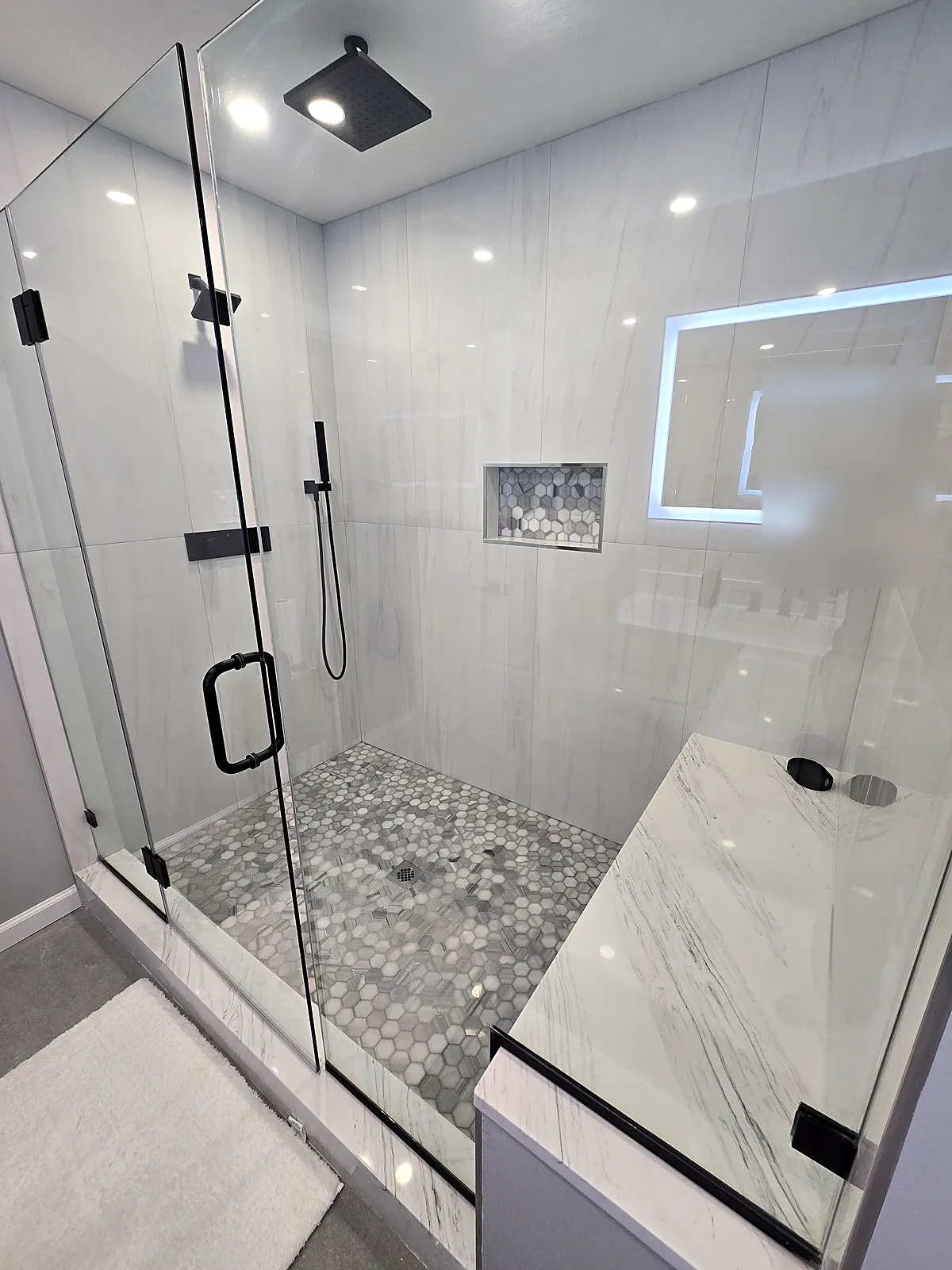 Bathroom remodel in Old Bethpage, NY: a frameless glass shower with a bench, a black rain head, a hex tile floor and a lighted niche wall
