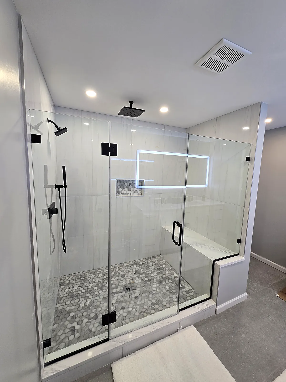 Bathroom remodel in Old Bethpage, NY: a large frameless glass shower with black fixtures, a built-in bench and a gray hex tile floor