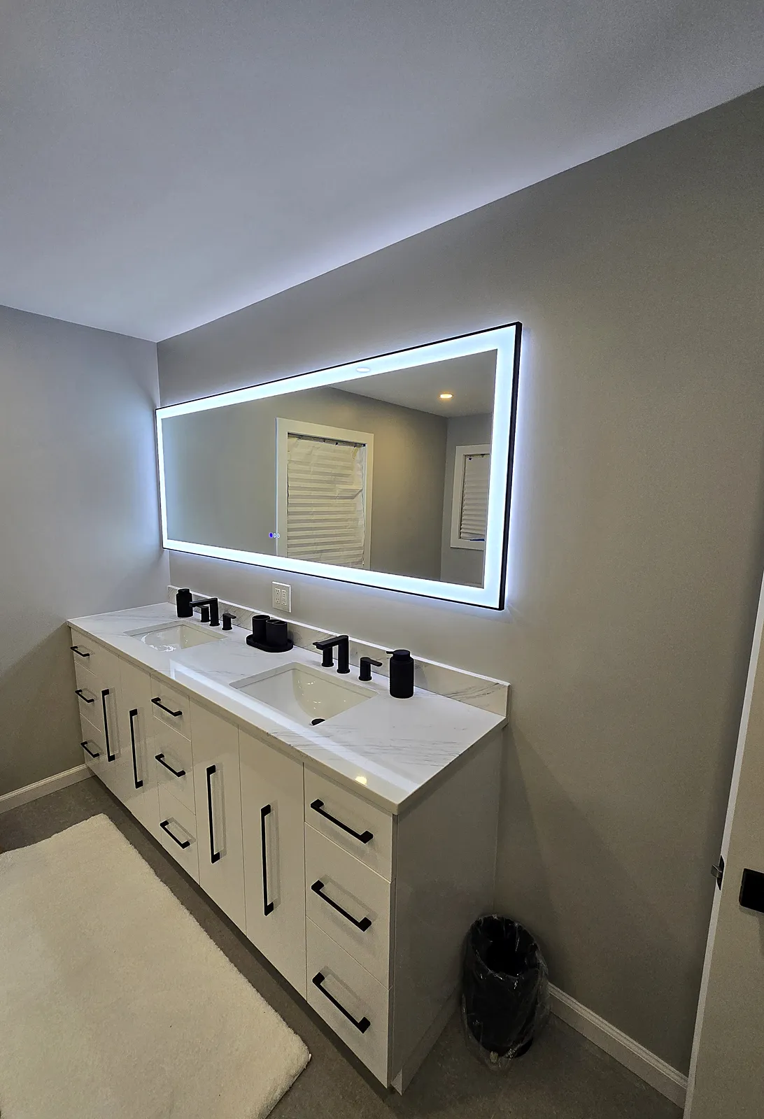 Bathroom remodel in Old Bethpage, NY: a white double vanity with black pulls and faucets under a long backlit mirror