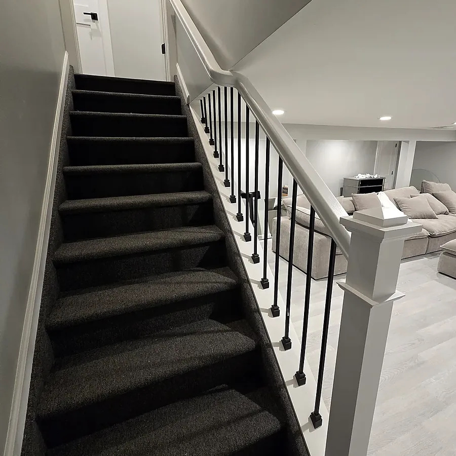 Basement staircase remodel in Old Bethpage, NY: a closed carpeted staircase with a bare wood edge replaced by a white railing with black metal spindles