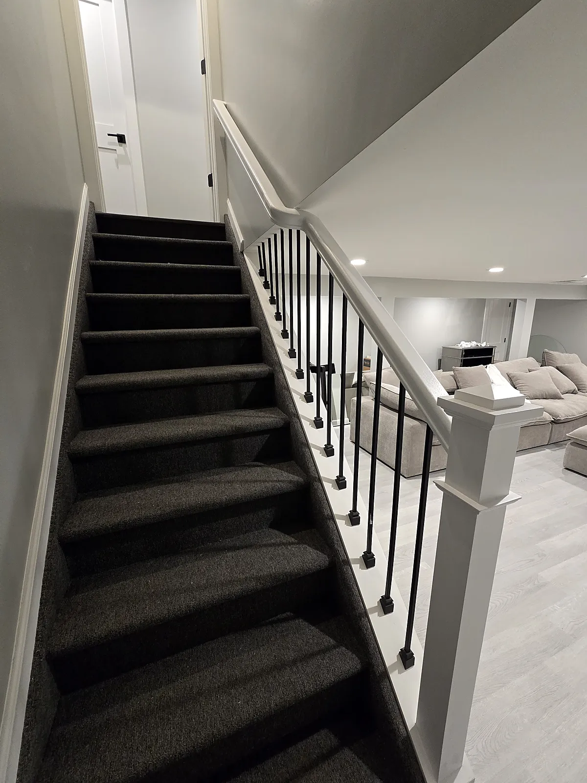 Basement staircase remodel in Old Bethpage, NY: carpeted basement stairs with a white railing, a square newel post and black metal spindles