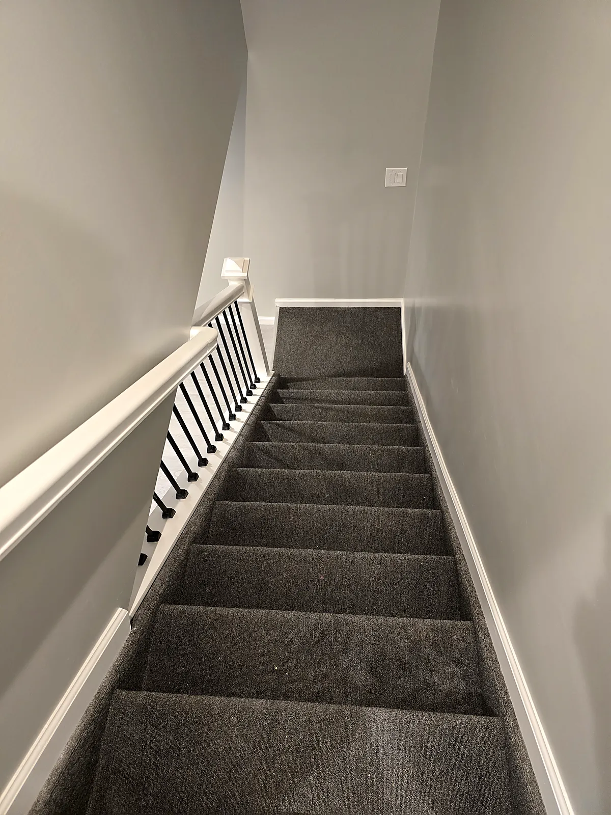 Basement staircase remodel in Old Bethpage, NY: a view down carpeted stairs with a white handrail and black metal spindles along one side