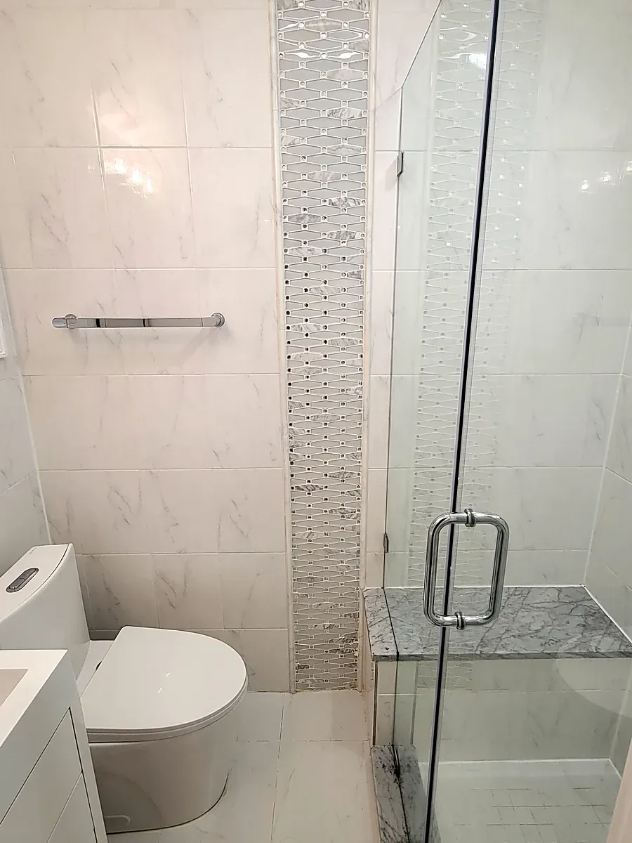 Bathroom remodel in Plainview, NY: white marble-look wall tile with a mosaic strip, glass shower door and marble bench