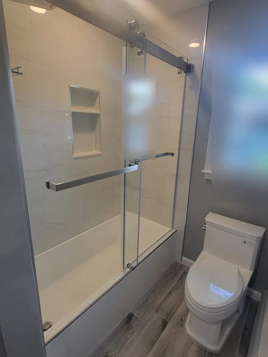 Bathroom remodel in Port Washington, NY: tub with sliding glass doors, tiled niche, gray walls and gray plank-look floor