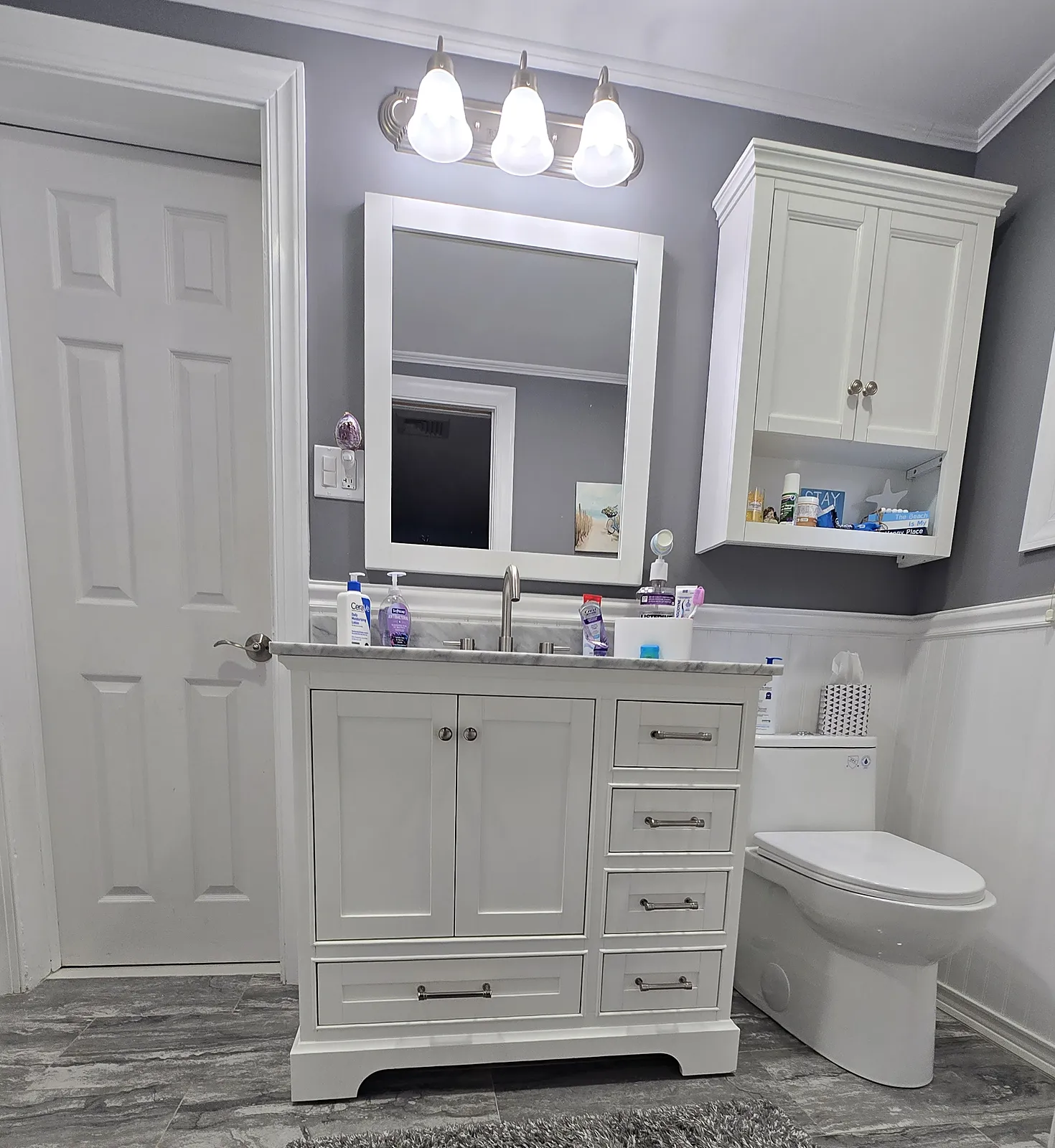 Bathroom remodel in Seaford, NY: white shaker vanity with a marble-look top, a white wall cabinet, wainscoting and gray stone-look floor tile