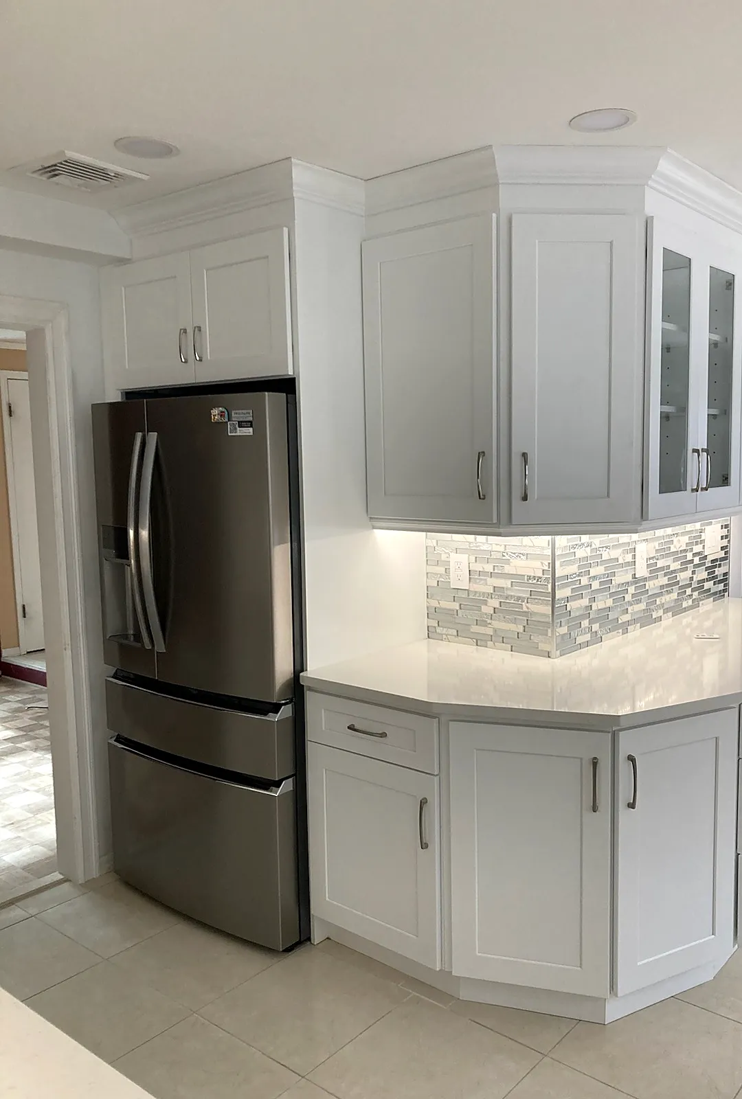 Kitchen remodel in Smithtown, NY: an angled corner of white shaker cabinets with glass doors, light counters and a stainless fridge