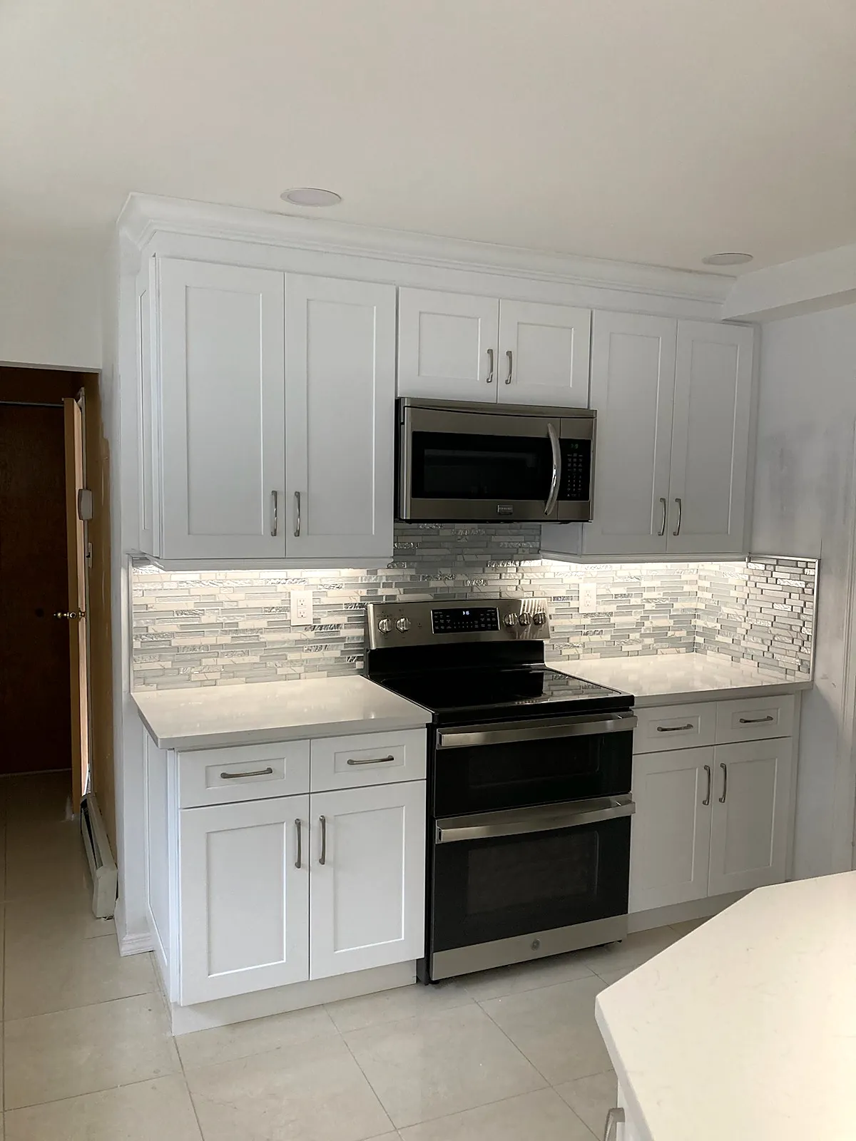 Kitchen remodel in Smithtown, NY: a white shaker cabinet wall with a stainless range, microwave and lit glass mosaic backsplash