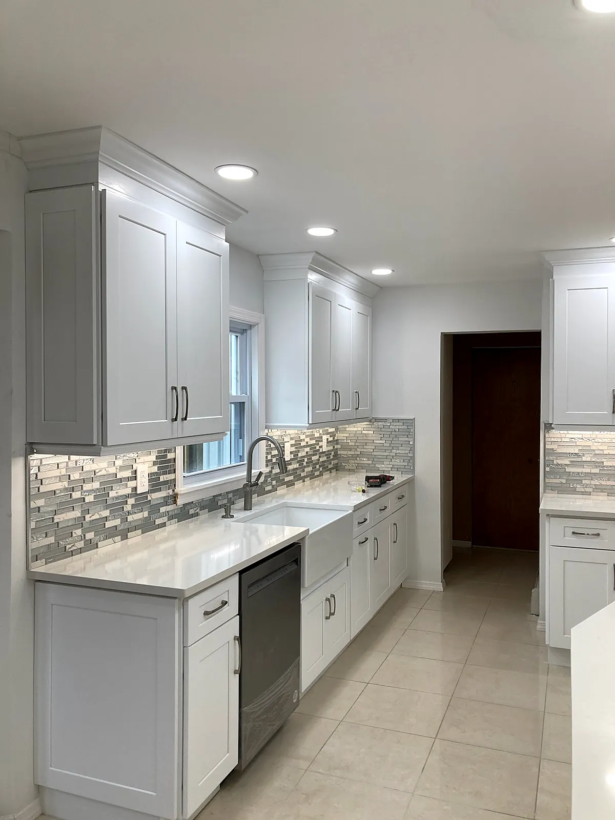 Kitchen remodel in Smithtown, NY: a galley run of white shaker cabinets with a farmhouse sink, dishwasher and glass mosaic backsplash