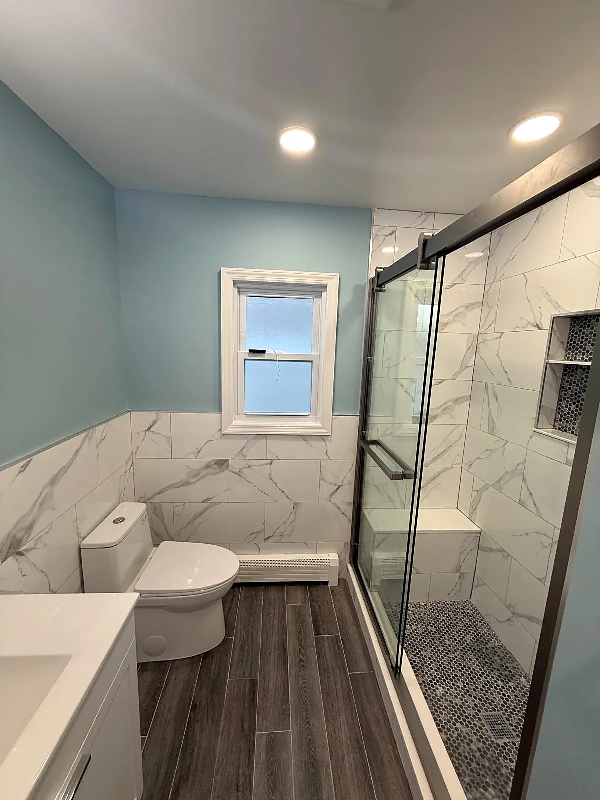 Bathroom remodel in Wantagh, NY: a sliding glass shower with a bench, niche and pebble floor, marble-look wainscot tile and light blue walls